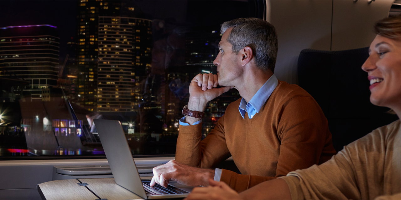 Middle-aged couple on a train at nighttime. Male is looking thoughtfully out the window.